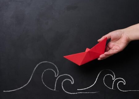 A female hand holds a red paper boat, with chalk-drawn waves below, representing the concept of mentorship and business support