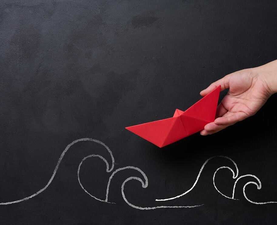 A female hand holds a red paper boat, with chalk-drawn waves below, representing the concept of mentorship and business support